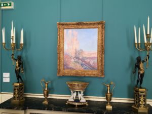 A Sisley painting above some ornate candelabras and pitchers.