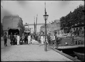 Circa 1930: On the right the Voordam and on the left Waagplein. On Waagplein some lorries of forwarding company P. Verwer. Near the lorries some cheese carriers and policemen. In the water of the Voordam are cargo ships.